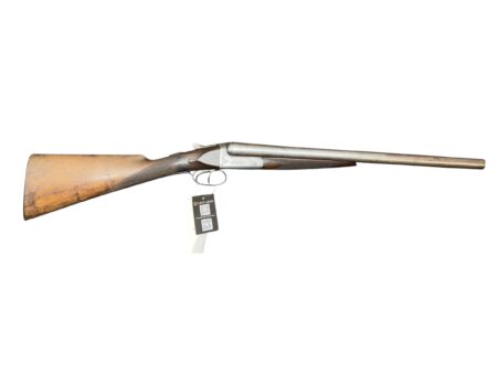 Side view of a vintage double-barreled shotgun with wooden stock and long metal barrels, tag hanging from trigger guard.