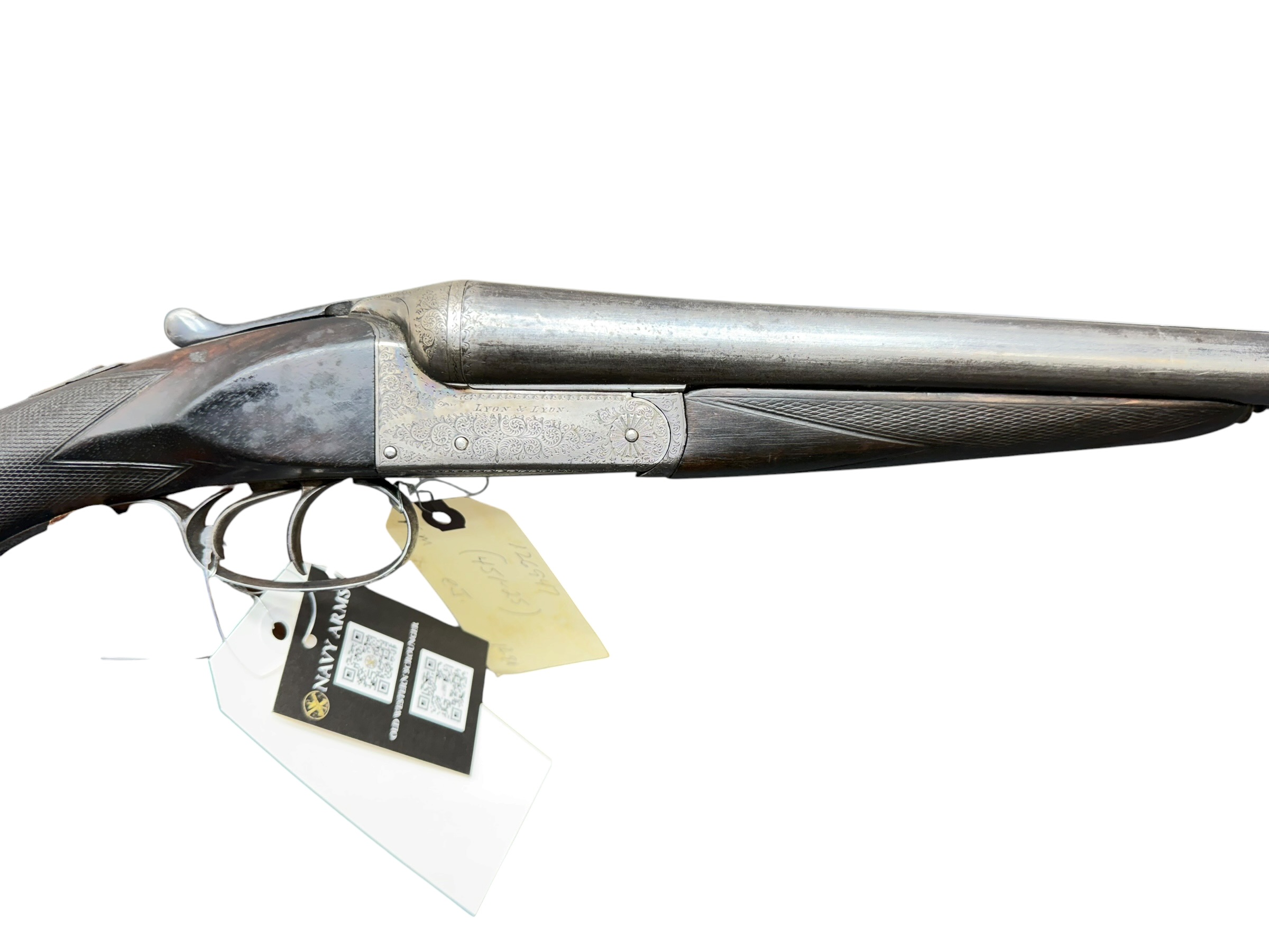 IMG_5380-Photoroom - OWS-Ammo Close-up of a decorative, engraved double-barrel shotgun showing the metal receiver, trigger guard, and long barrel with a hanging tag attached.